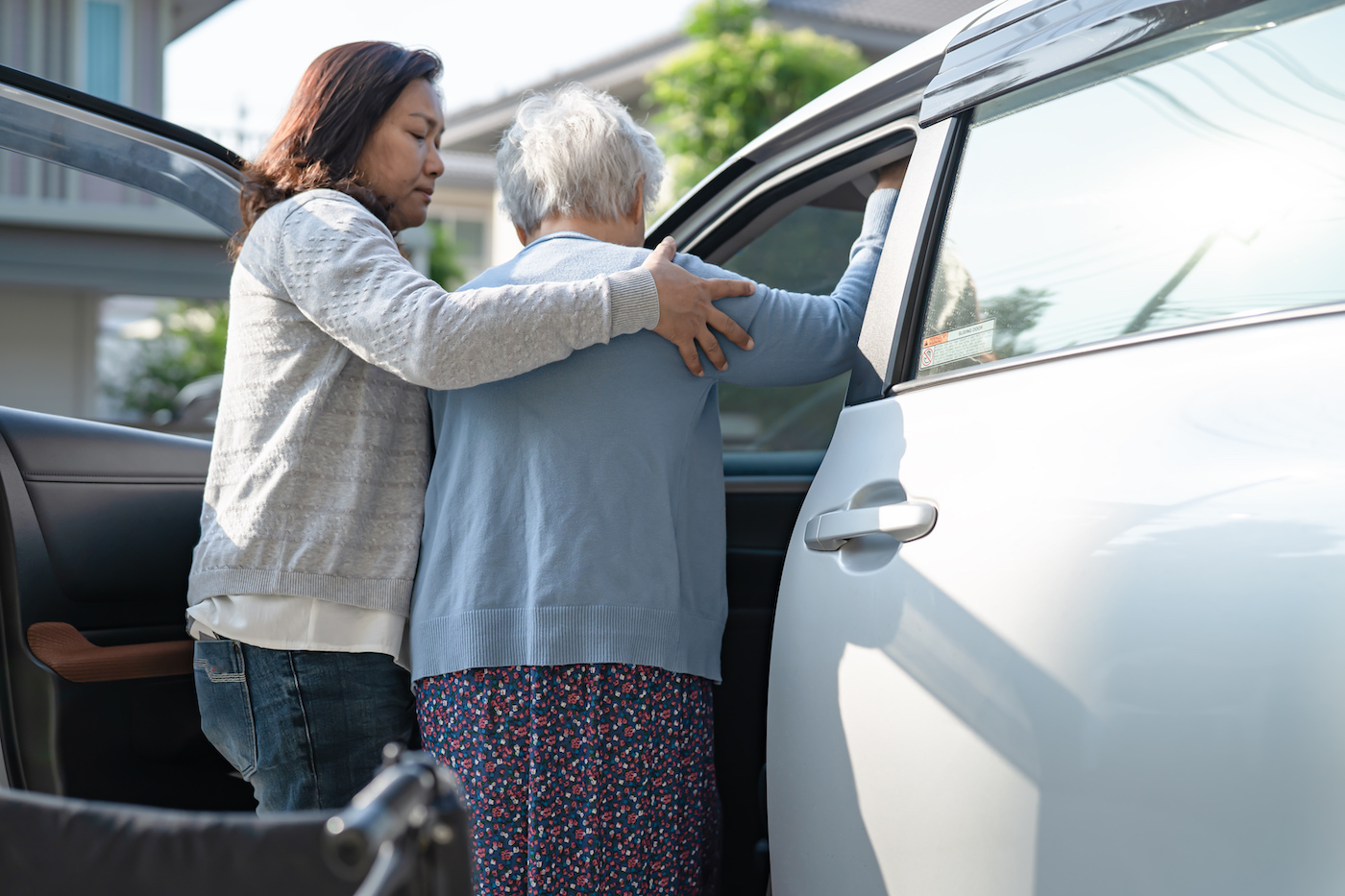 Strahlentherapiefahrten mit Stegersbach Taxi im Bezirk Oberwart und Bezirk Güssing. Junge charmante Taxifahrerin hilft eine ältere Frau aus dem Rollstuhl in das graue Taxi, bei den die Autotür offen ist, zu steigen um mit diesen von der Strahlentherapie nach Hause zu fahren.