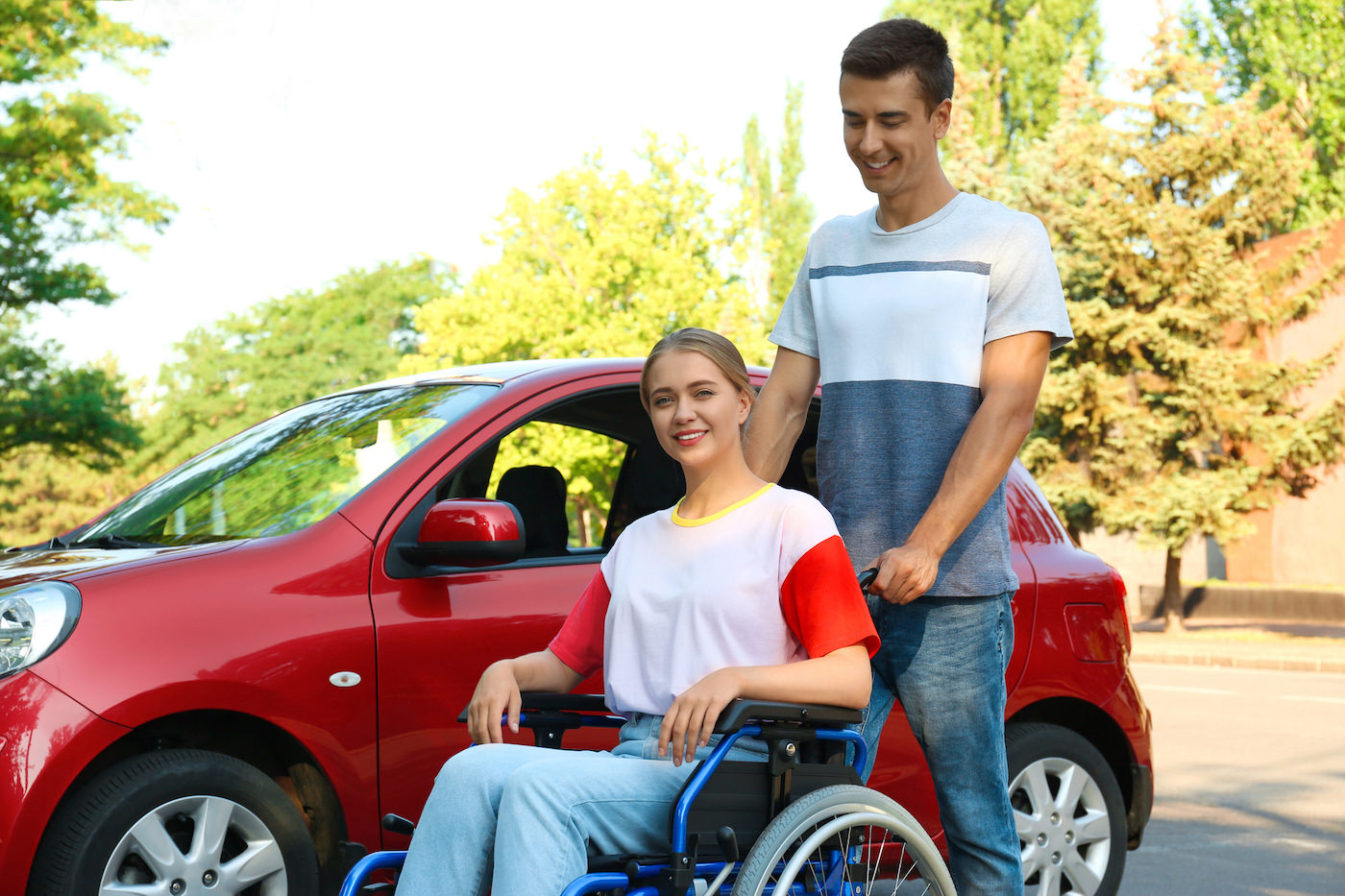 Rehabfahrten im Bezirk Oberwart und Bezirk Güssing Junger Taxifahrer schiebt Junge sympathische Frau nach dem sie mit eine roten Taxi gefahren sind an einen Ort mit Bäumen im Hintergrund zu sehen zur Rehab.